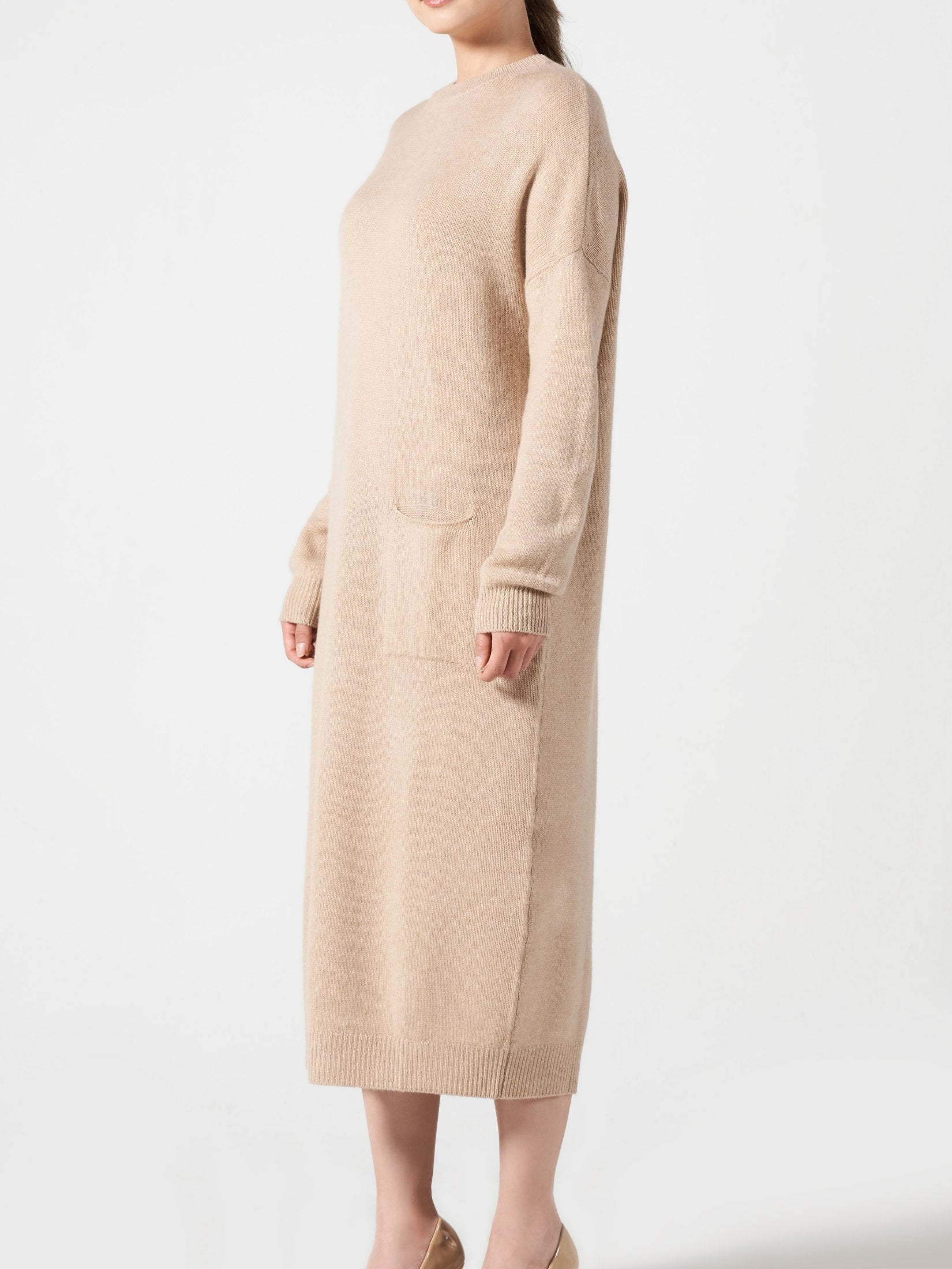 Woman wearing a camel-colored loose fit cashmere dress with one pocket and long sleeves against white background