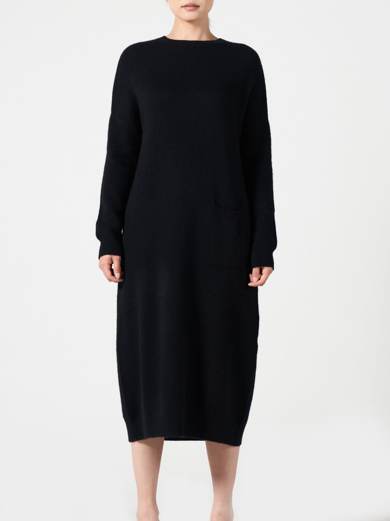 Woman wearing a black cashmere loose fit dress with one pocket and round neck standing against white background