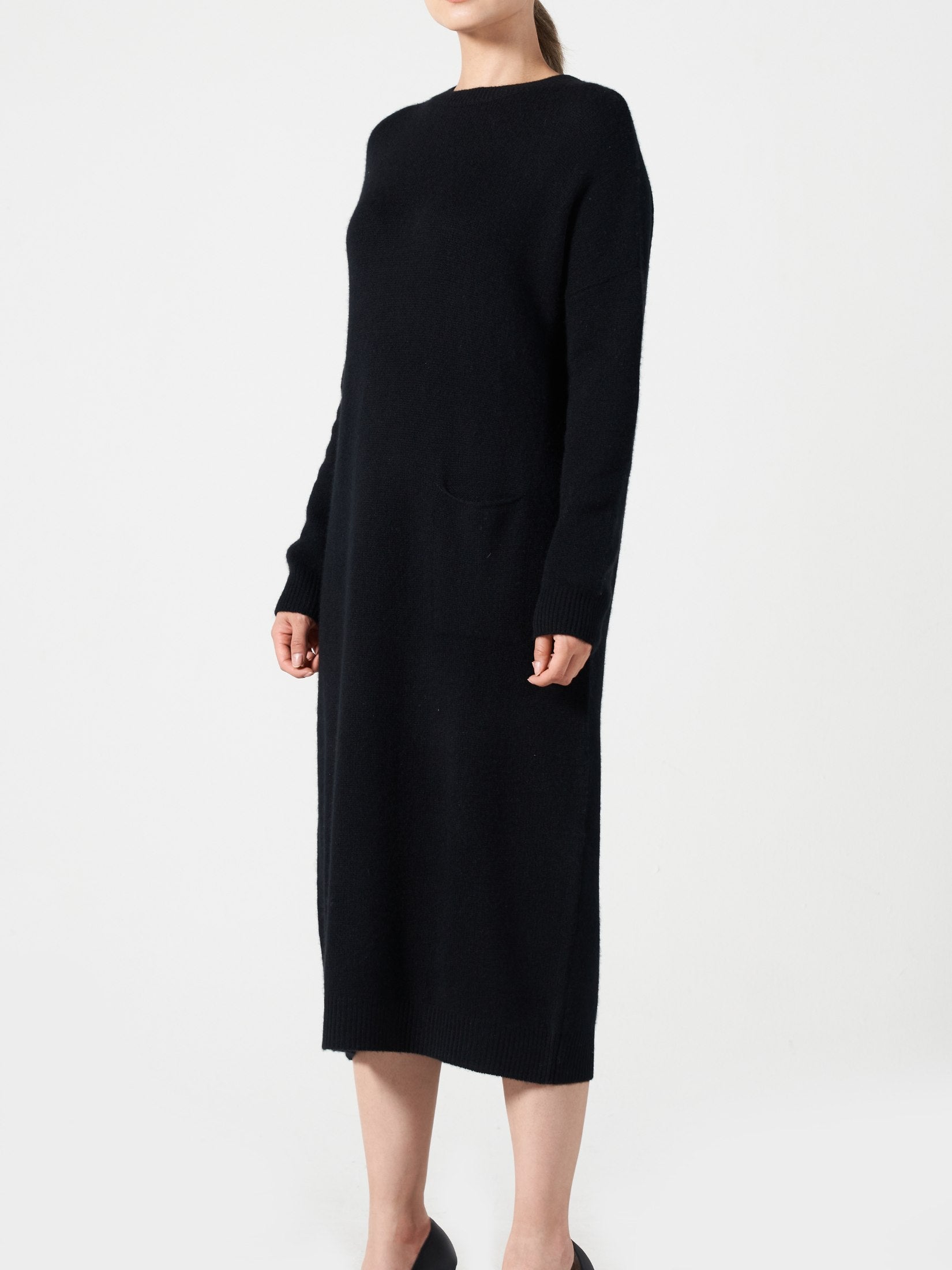 Woman wearing a black loose fit cashmere dress with one pocket and long sleeves on a white background
