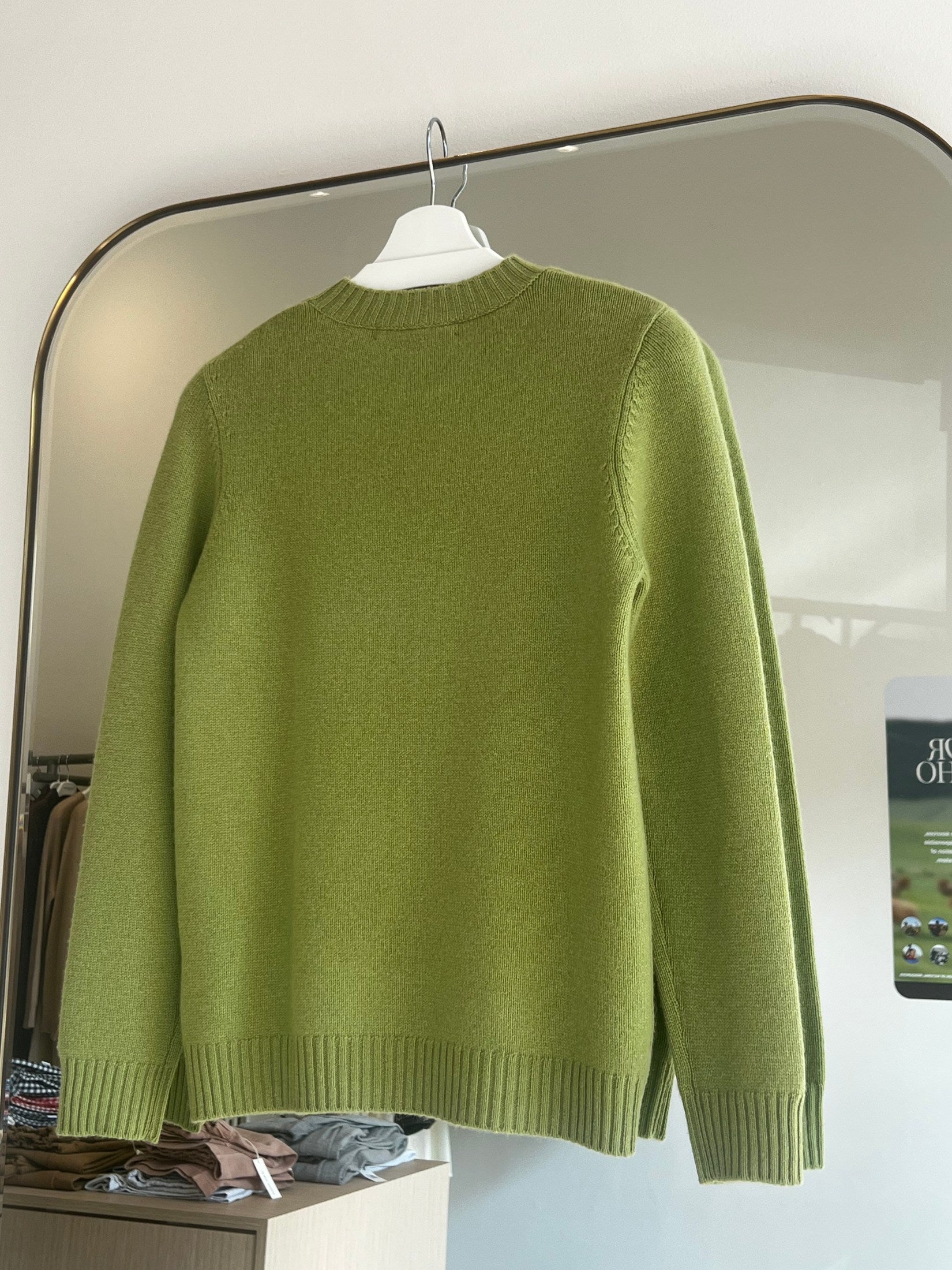 Green round neck cashmere sweater hanging on a white hanger in front of a large mirror.