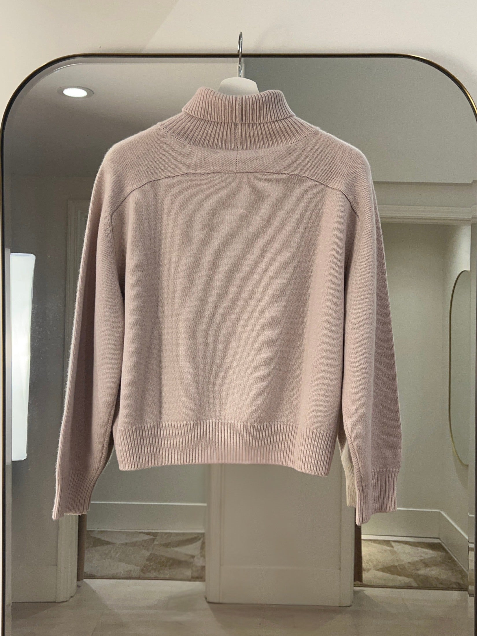 Light pink crop length turtle neck cashmere sweater with raglan shoulders hanging on a hanger in a room.