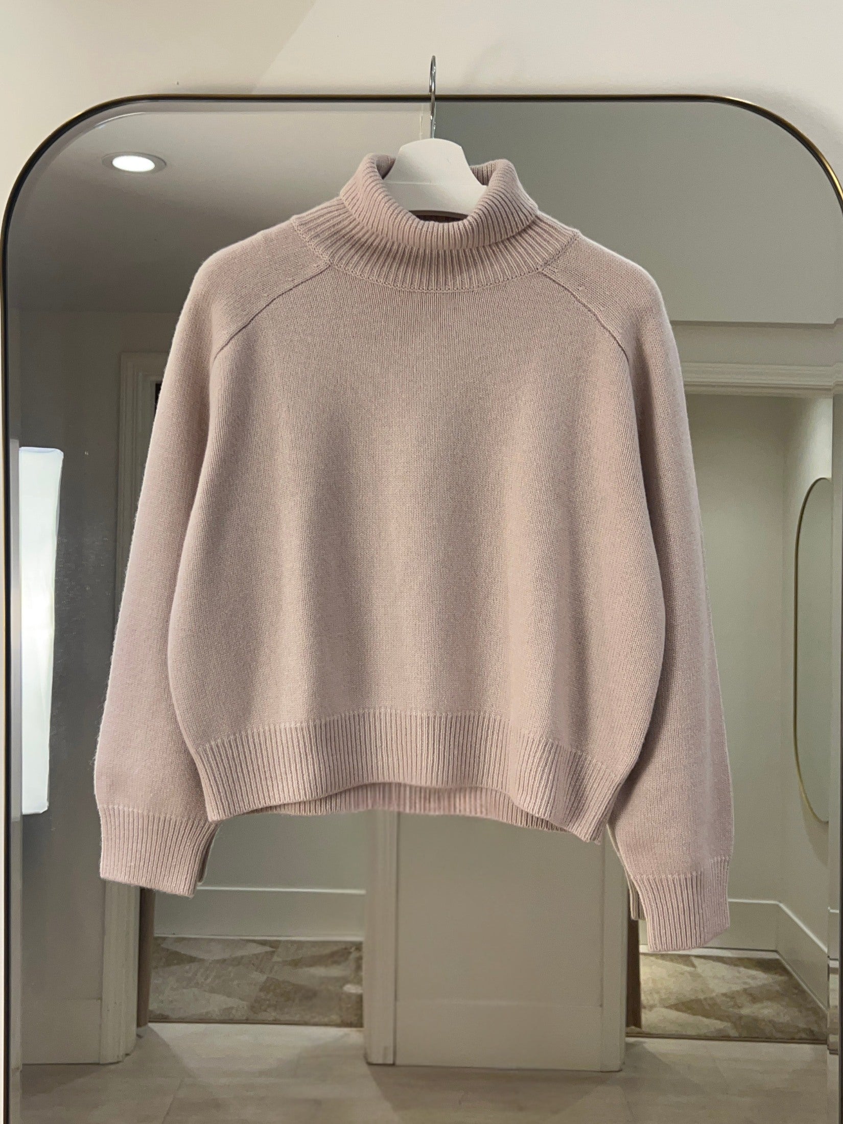 Light pink crop length turtle neck cashmere sweater with raglan shoulders hanging on a white hanger in a room