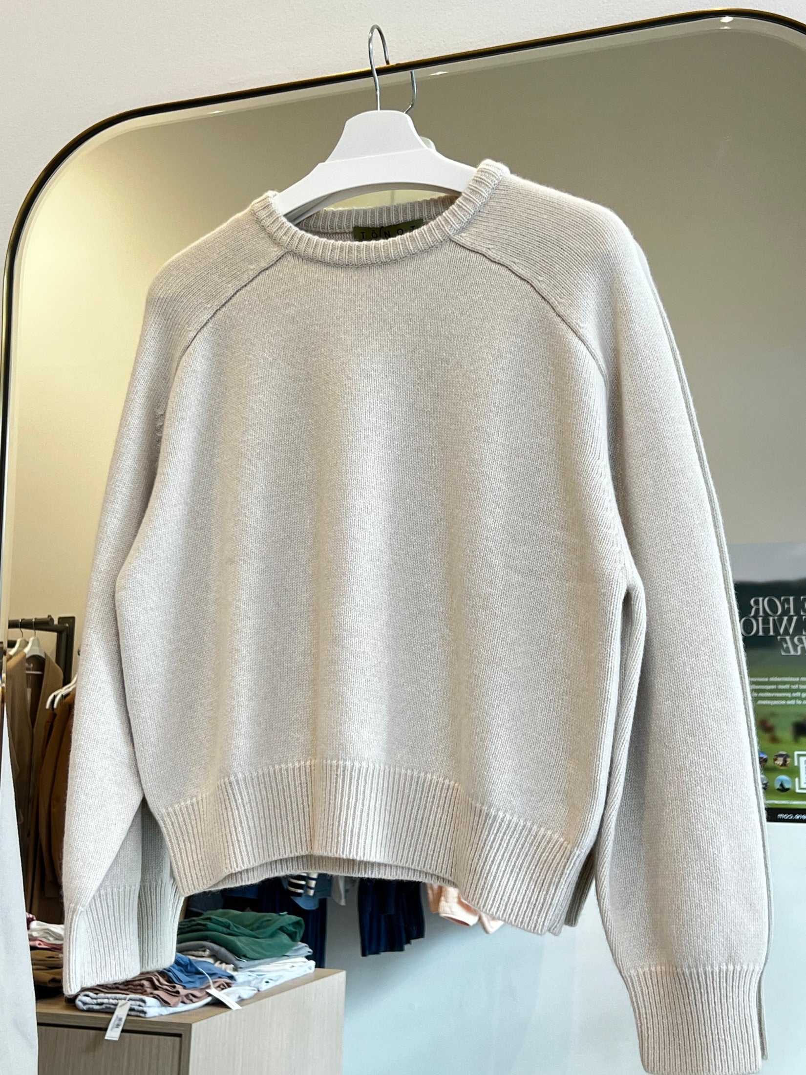 Beige round neck cropped cashmere sweater with raglan sleeves hanging on a white hanger in a store.