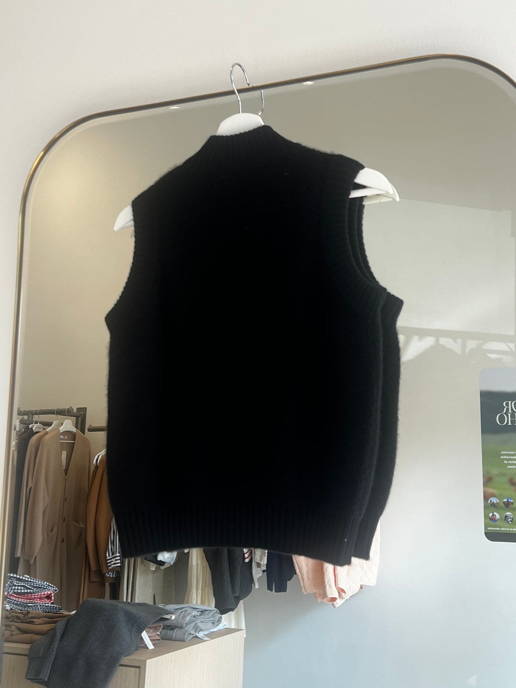 Black moss stitch cashmere button vest hanging on a white hanger in front of a mirror.