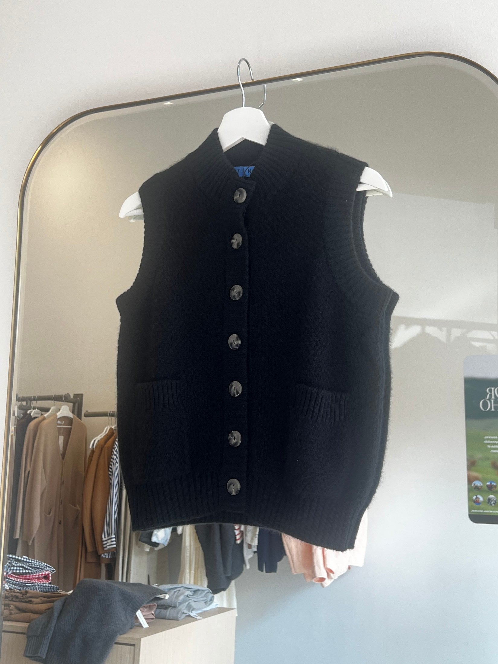 Black moss stitch cashmere button vest with two front pockets hanging on white hanger against mirror background