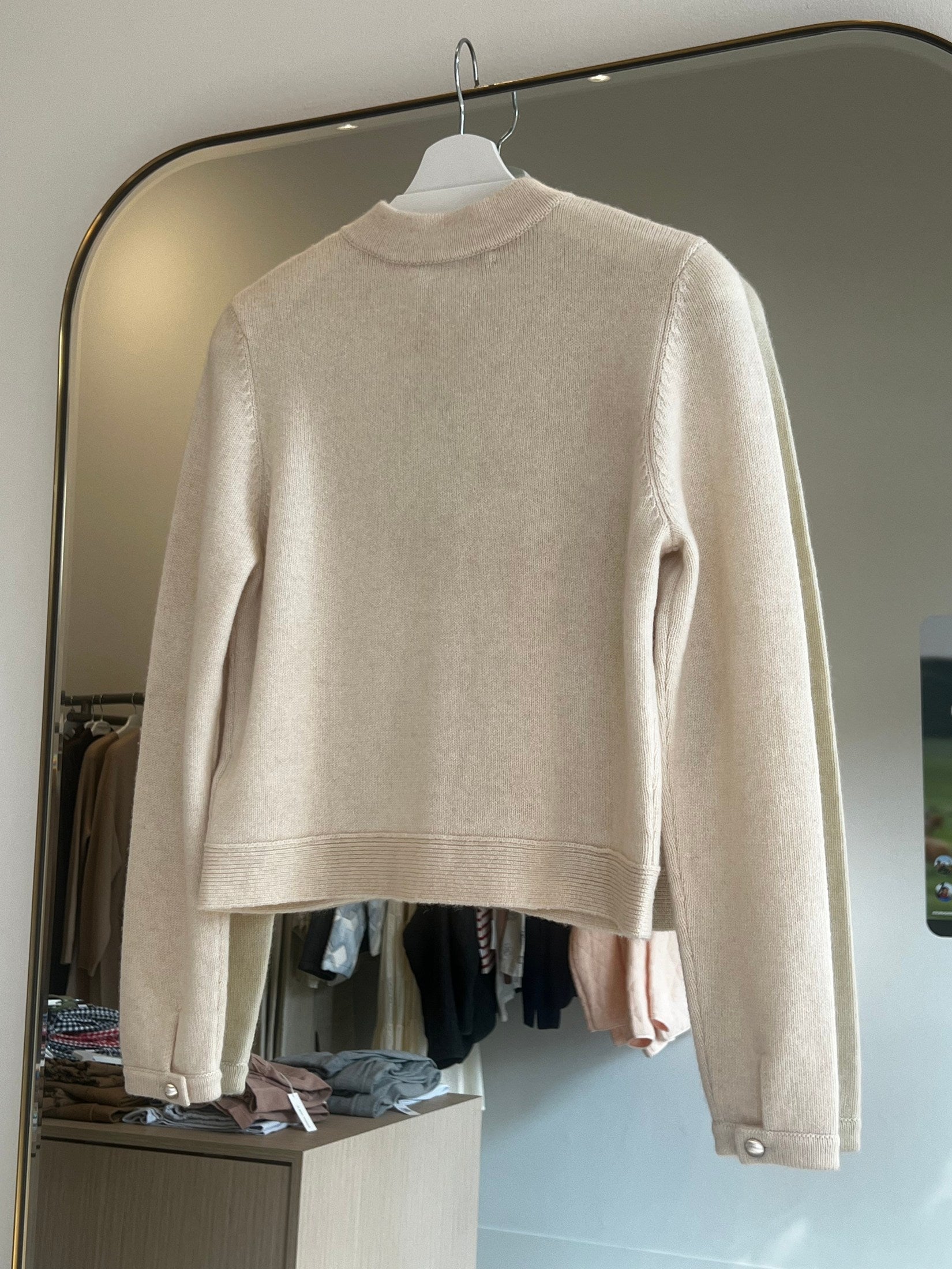 Beige crop-length cashmere cardigan with long sleeves hanging on a white hanger in front of a mirror