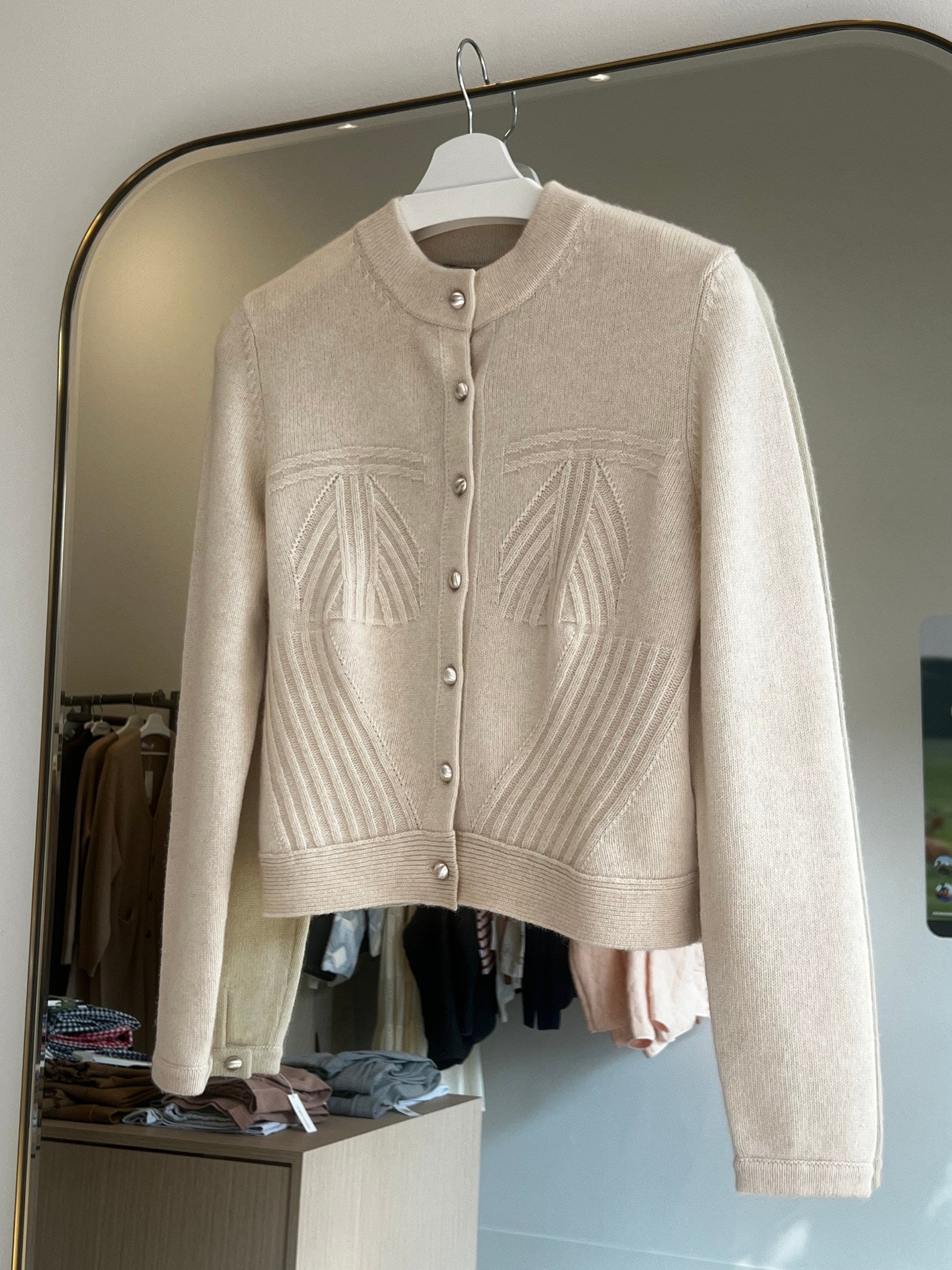 Beige crop-length cashmere cardigan with button closure and textured stitch design hanging on white hanger.