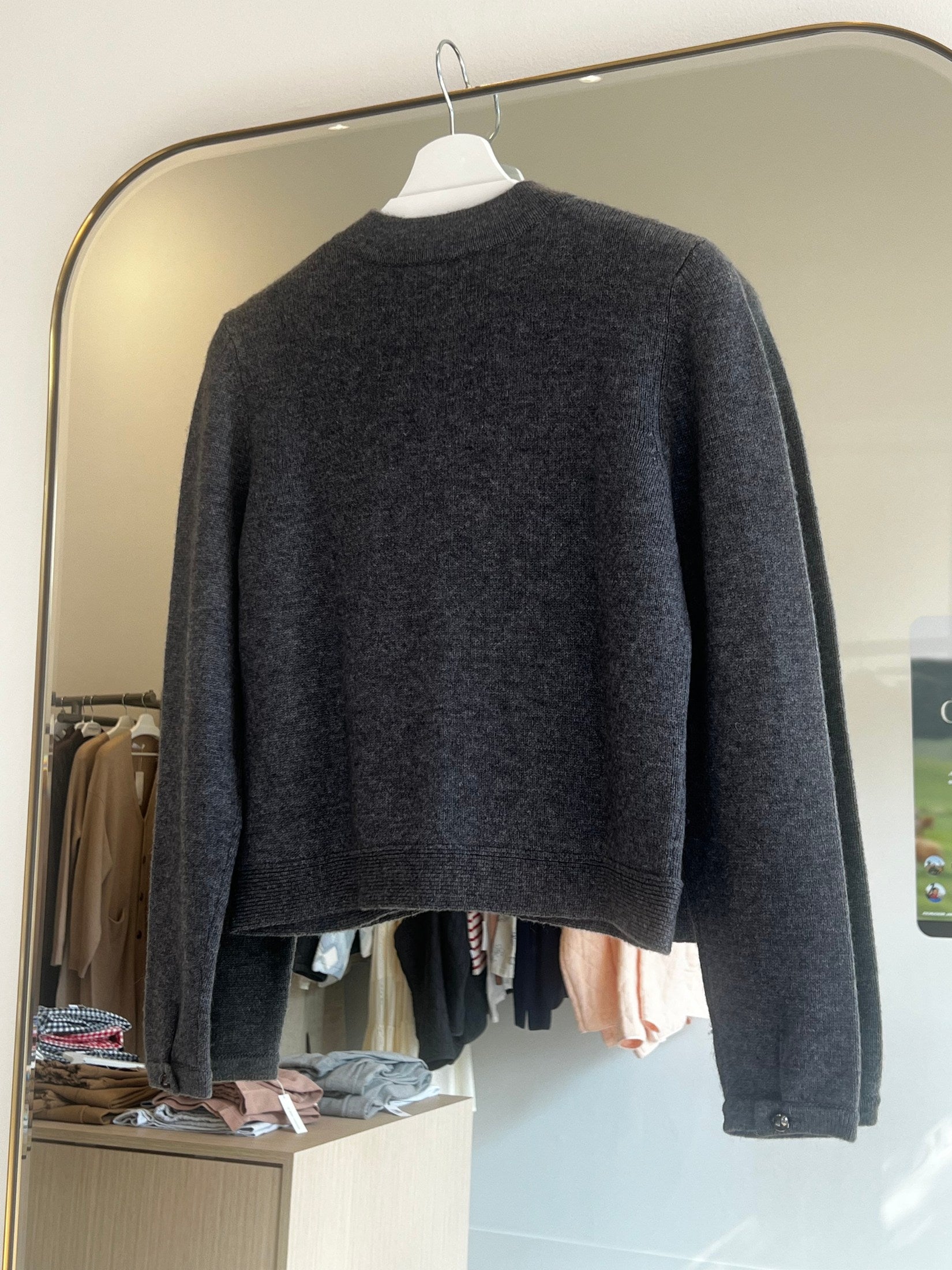 Dark gray shift stitch crop cashmere cardigan hanging on white hanger in boutique setting with clothing racks.